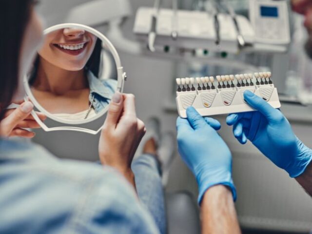 implant sonrası iyileşme rehberi Diş implantı sonrası aynada gülümseyerek dişlerini kontrol eden kadın ve renk skalası tutan diş hekimi.