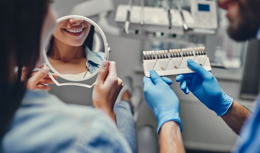 Diş implantı sonrası aynada gülümseyerek dişlerini kontrol eden kadın ve renk skalası tutan diş hekimi.